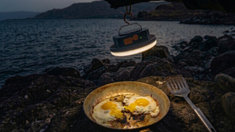 Cooking eggs on a pan with a camping light above, set against a natural backdrop.