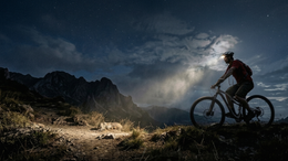 Person riding a bicycle on a mountain trail with a dramatic sky and light source.
