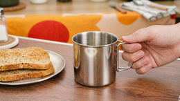 Hand holding a stainless steel mug on a wooden table with toast and a plate in the background.