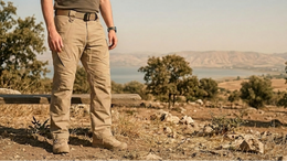 Person standing outdoors in a natural landscape with mountains and water in the background.